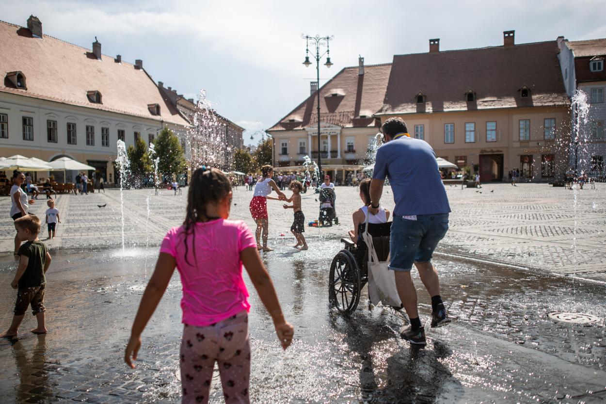 The image shows one of the teams experiencing a pavement-level fountain in Sibiu, Romania. People of different ages are present, including children playing in the fountain and a wheelchair user pushed by somebody else.