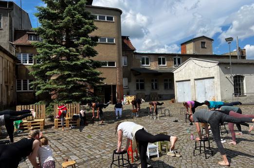 A group of people doing chair yoga in a cobblestone courtyard on a sunny day, with an industrial building and a large tree in the background