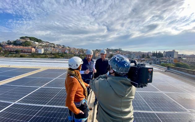 A group of four people on a rooftop covered with solar panels. The setting appears to be an urban area, with buildings and greenery visible in the background under a partly cloudy sky.